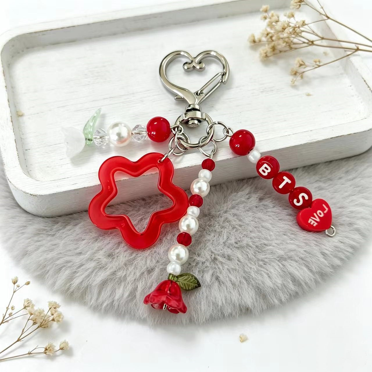 A decorative keychain with a silver heart-shaped clasp, red beads, a red flower-shaped charm, white pearl-like beads, a red rose charm, and red letter beads spelling BTS and LOVE, displayed on a white tray over a gray faux-fur surface.