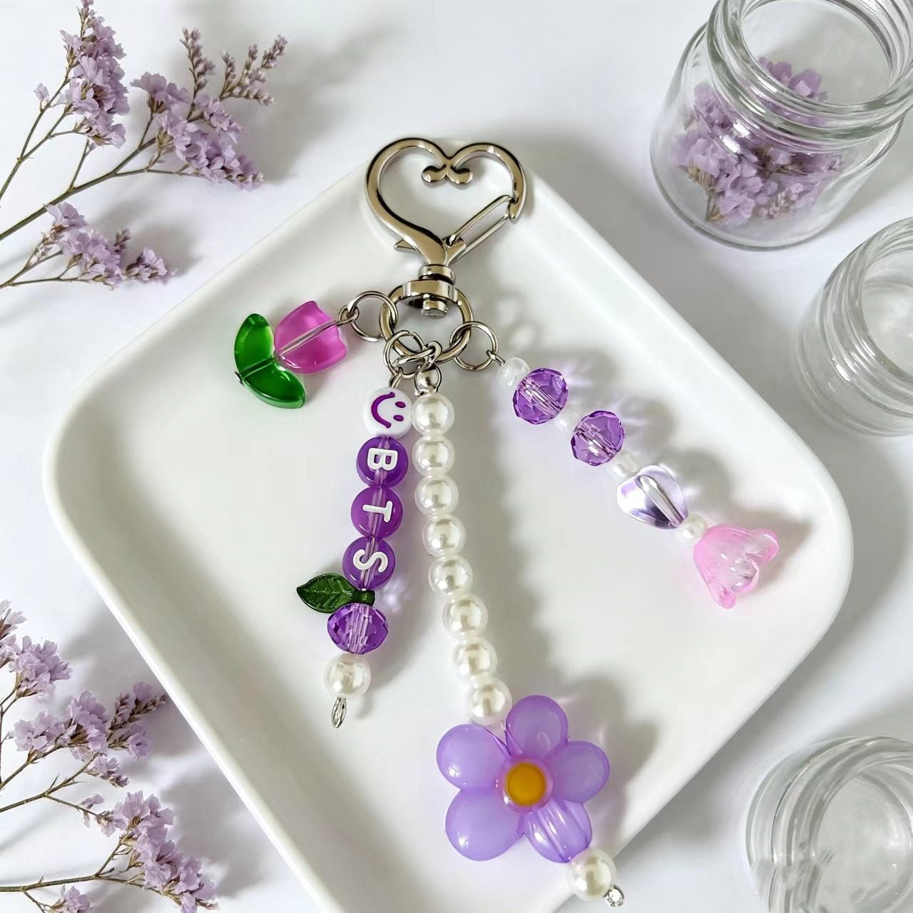A decorative keychain with a silver heart-shaped clasp, purple letter beads spelling 'CUBIS', a dangling pearl bead strand, a purple flower charm, and small purple and pink beads, displayed on a white tray with dried flowers and glass jars around it.