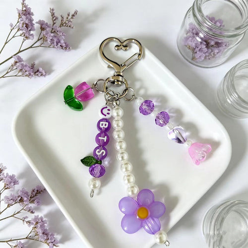 A decorative keychain with a silver heart-shaped clasp, purple letter beads spelling 'CUBIS', a dangling pearl bead strand, a purple flower charm, and small purple and pink beads, displayed on a white tray with dried flowers and glass jars around it.