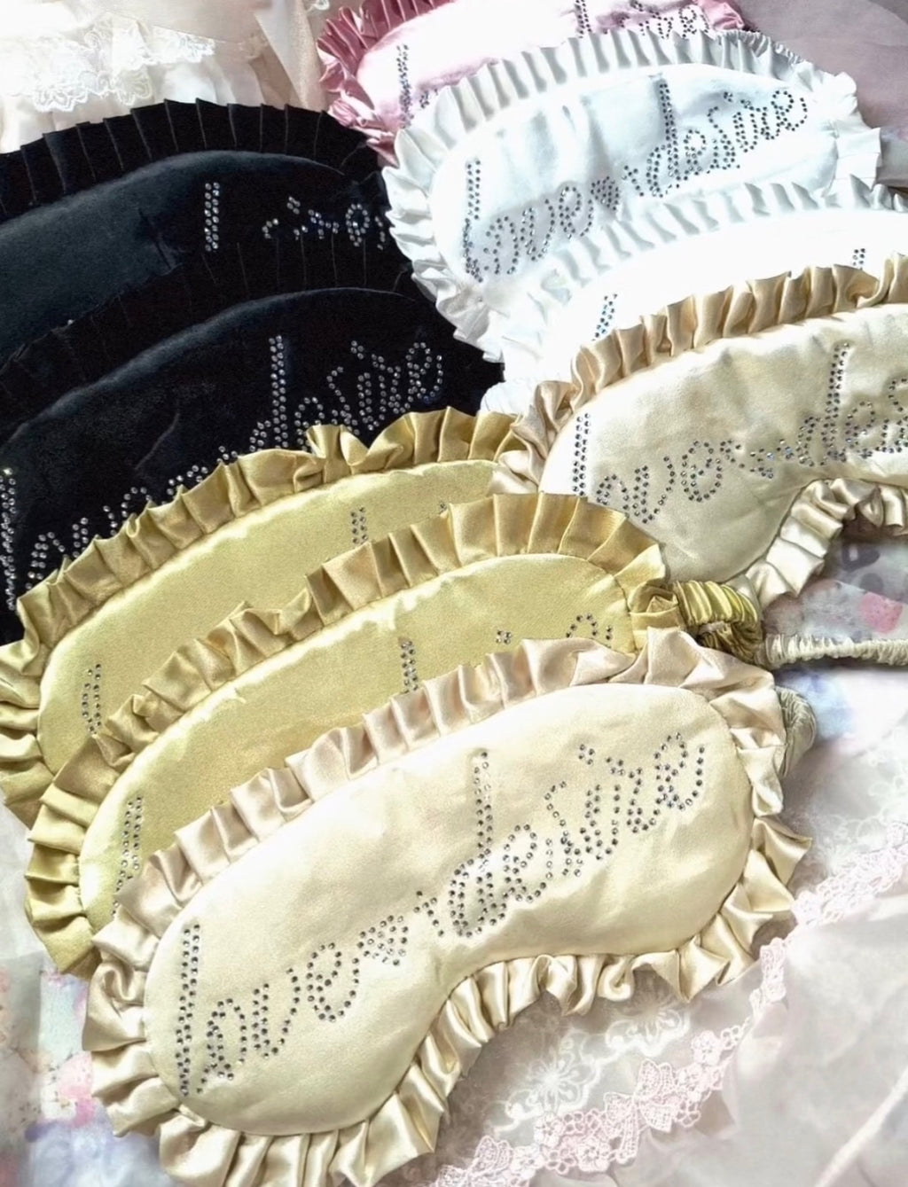 A set of satin sleep masks with ruffled edges and rhinestone lettering, shown in black, white, and champagne beige, with the word 'love' spelled out in rhinestones.