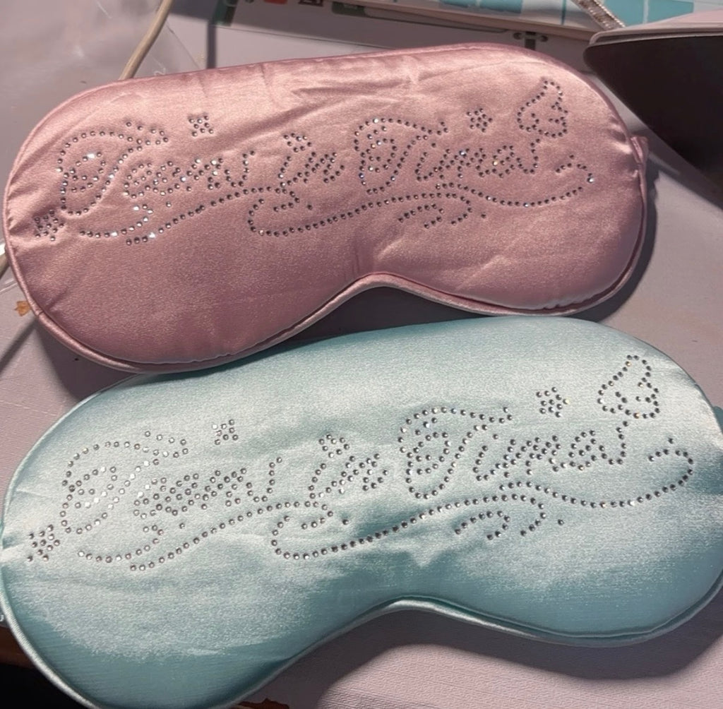Two satin sleep masks, one light pink and one light blue, both featuring rhinestone-embellished cursive text reading 'Sleep In Time' on a light-colored surface.