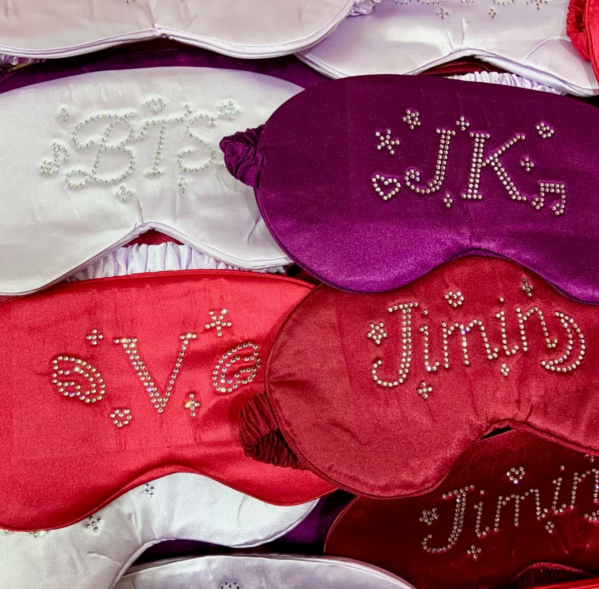 A close-up of multiple satin sleep masks in white, purple, red, and burgundy, each decorated with rhinestone monogram lettering.