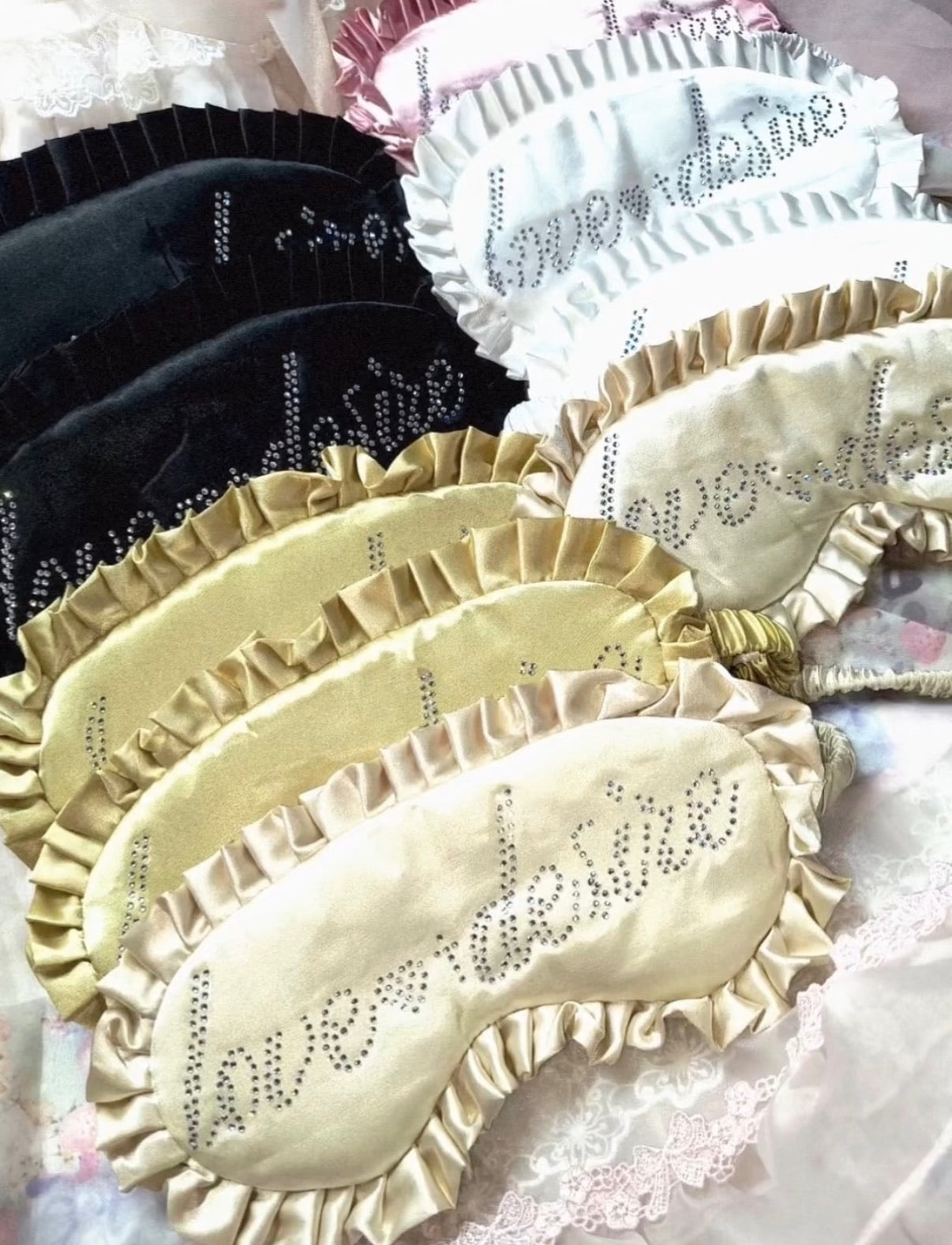 A set of satin sleep masks with ruffled edges and rhinestone lettering, shown in black, white, and champagne beige, with the word 'love' spelled out in rhinestones.